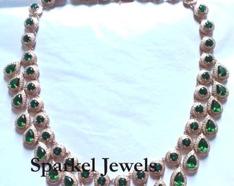 Op de afbeelding: Een gouden ketting met een dubbele rij smaragdgroene peervormige stenen en een hangende rij smaragdgroene peervormige stenen. De ketting is versierd met kleinere heldere stenen. De ketting is gelabeld met "Sparkel Jewels".