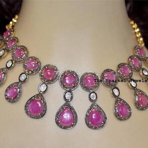 Puede incluir: Un collar con piedras cabujón rosas redondas y en forma de pera, engastadas en marcos de metal oscuro con pequeñas piedras claras. El diseño incluye una fila central de piedras redondas con gotas colgantes. El texto dice The Sparkel Jewels.