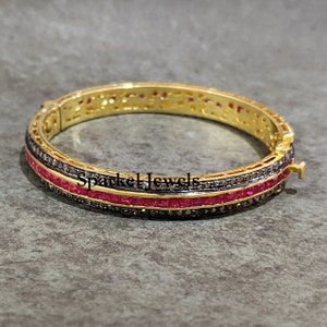 May include: Gold-toned bangle bracelet with a row of red gemstones and a row of small, dark gemstones. The bracelet has the text "Sparkel Jewels" on it.