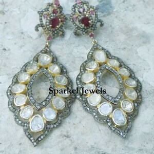 May include: A pair of silver dangle earrings with a floral design and a large ruby gemstone at the top. The earrings are adorned with smaller diamonds and have a teardrop shape.