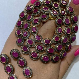 May include: A gold necklace and matching earrings set. The jewellery features oval, deep red gemstones surrounded by a halo of smaller, clear stones. The gemstones are set in a gold-coloured metal.