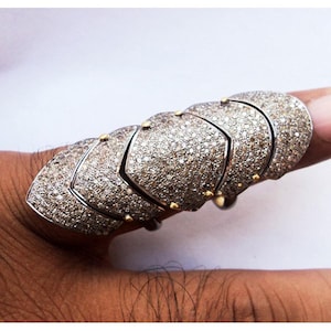 May include: A full-finger armour ring featuring five articulated, overlapping silver-toned segments. Each segment is densely encrusted with small, shimmering white stones and accented with tiny, polished gold-toned metal studs at the joints.