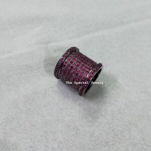 Puede incluir: Un anillo de banda ancha con un acabado de metal oscuro y múltiples filas de pequeñas piedras de color rosa vibrante que cubren la superficie. El texto The Sparkel Jewels está impreso en blanco en el lateral del anillo, de forma cilíndrica.