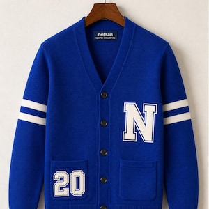 Pode incluir: Um cardigan azul royal com detalhes brancos. O suéter apresenta um grande "N" branco na frente, junto com o número "20". Listras brancas adornam as mangas. A marca "nersan sports industries" é visível em uma etiqueta.