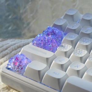 May include: A white mechanical keyboard featuring custom artisan keycaps with a translucent, textured purple and blue crystalline design. The keycaps are positioned on the left side of the keyboard, replacing standard keys, with a smooth white finish.