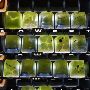 Puede incluir: Primer plano de un teclado con teclas personalizadas. Las teclas están hechas de resina verde translúcida con diseños de tinta negra y blanca. Las teclas están dispuestas en filas y columnas, y el teclado está iluminado desde abajo, creando un efecto brillante.