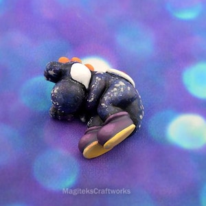 Puede incluir: Figura artesanal de arcilla polimérica de un personaje, con cuerpo azul oscuro y detalles blancos y naranjas. La figura está tumbada, con zapatos morados y suelas amarillas. Fondo morado borroso con bokeh.