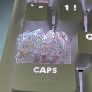 May include: Iridescent crystal keycaps on a computer keyboard. The keycaps are shaped like clusters of crystals and sit on a light green keyboard. The "CAPS" keycap has the text "MagiteksCraftworks" below it.