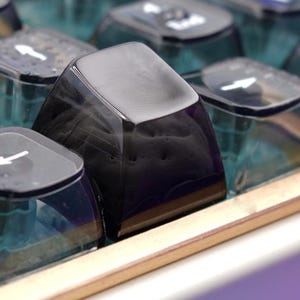 May include: A mechanical keyboard featuring translucent dark blue keycaps with white lettering, including Enter, Shift, and Ctrl keys. A single solid black keycap is positioned in the center. The keyboard has a gold-colored frame and white base.