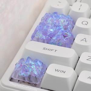 May include: A close-up of a white mechanical keyboard with custom keycaps. The keycaps are white, and the custom keycaps are iridescent purple crystal shapes. The keyboard has the text "SHIFT" and "WIN" on the keys.