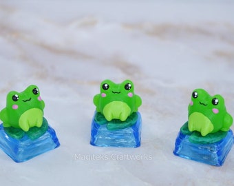 May include: Three green frog keycaps with blue and green bases. The frogs have black eyes and pink cheeks.