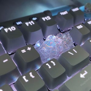 May include: Close-up of a computer keyboard with gray keycaps and white illuminated lettering. A clear, iridescent crystal sits on the keyboard. The keyboard keys include F9, F10, F11, F12, and other function keys.