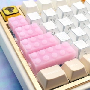 May include: A mechanical keyboard featuring white and beige keycaps, a gold-colored frame, and custom pink building block-style keys. A yellow keycap with a black graphic is visible in the top left, and the text MagiteksCraftworks is on the side.