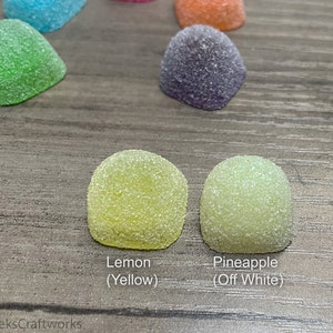Op de afbeelding: Close-up van zes verschillende gekleurde gummy snoepjes. De snoepjes zijn koepelvormig en hebben een korrelige textuur. De kleuren zijn groen, blauw, oranje, paars, geel en off-white. De snoepjes zijn gelabeld met hun smaken: Lemon (Geel), Pineapple (Off White).