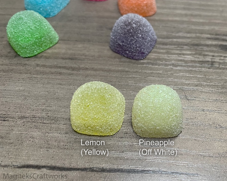 Op de afbeelding: Close-up van zes verschillende gekleurde gummy snoepjes. De snoepjes zijn koepelvormig en hebben een korrelige textuur. De kleuren zijn groen, blauw, oranje, paars, geel en off-white. De snoepjes zijn gelabeld met hun smaken: Lemon (Geel), Pineapple (Off White).