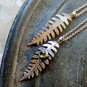 Pode incluir: Dois pingentes prateados em forma de folha de feto em correntes delicadas. Um pingente apresenta uma pequena gema verde redonda. Os pingentes são exibidos em uma superfície texturizada cinza escuro, com um fundo de cristais brancos.