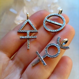 Könnte beinhalten: Drei silberne Anhänger mit astrologischen Symbolen: ein Dreieck mit einem Kreuz, ein Kreis mit einer Linie durch ihn und ein Kreis mit einer Mondsichel und einem Kreuz.