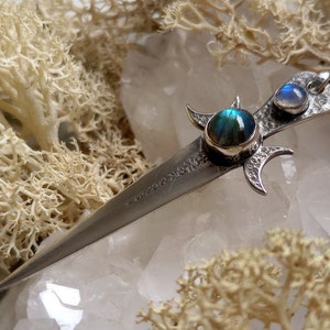 May include: A silver pendant in the shape of a dagger with two iridescent blue moonstone gems set into the hilt. The pendant is surrounded by white crystals.