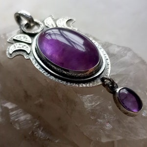 May include: A silver pendant with a large oval amethyst gemstone and a smaller amethyst dangle. The pendant features a detailed design with crescent moon shapes.