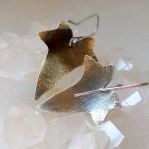 May include: A pair of silver and gold leaf earrings in the shape of a stylized leaf. The earrings are hanging from a white surface.
