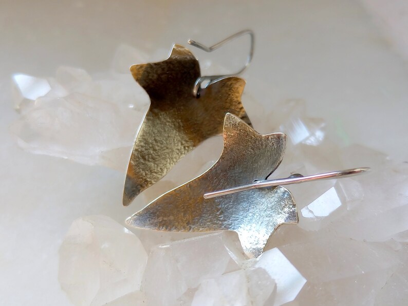 May include: A pair of silver and gold leaf earrings in the shape of a stylized leaf. The earrings are hanging from a white surface.
