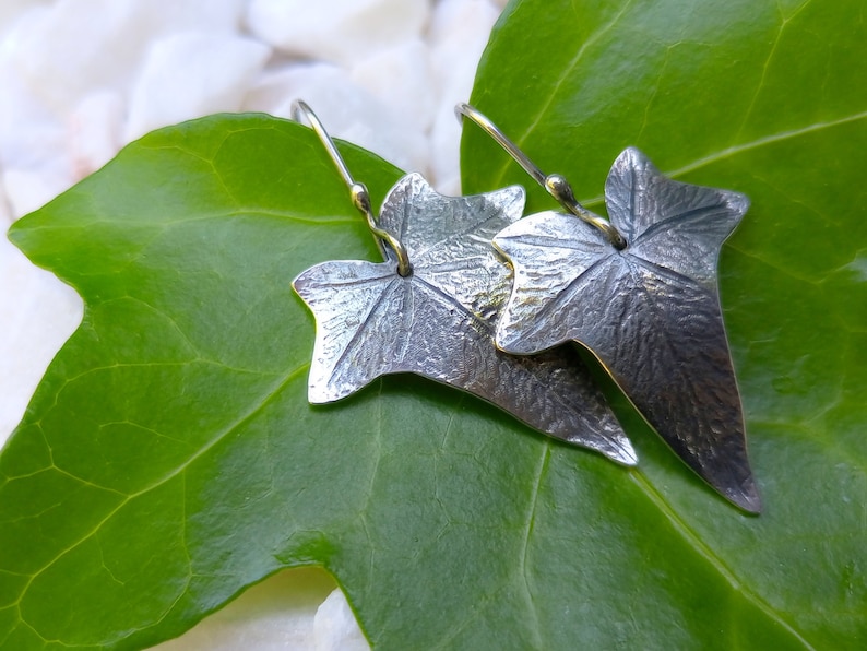 May include: A pair of silver earrings shaped like ivy leaves. The leaves are textured and have a realistic look. The earrings are hanging from a green leaf.