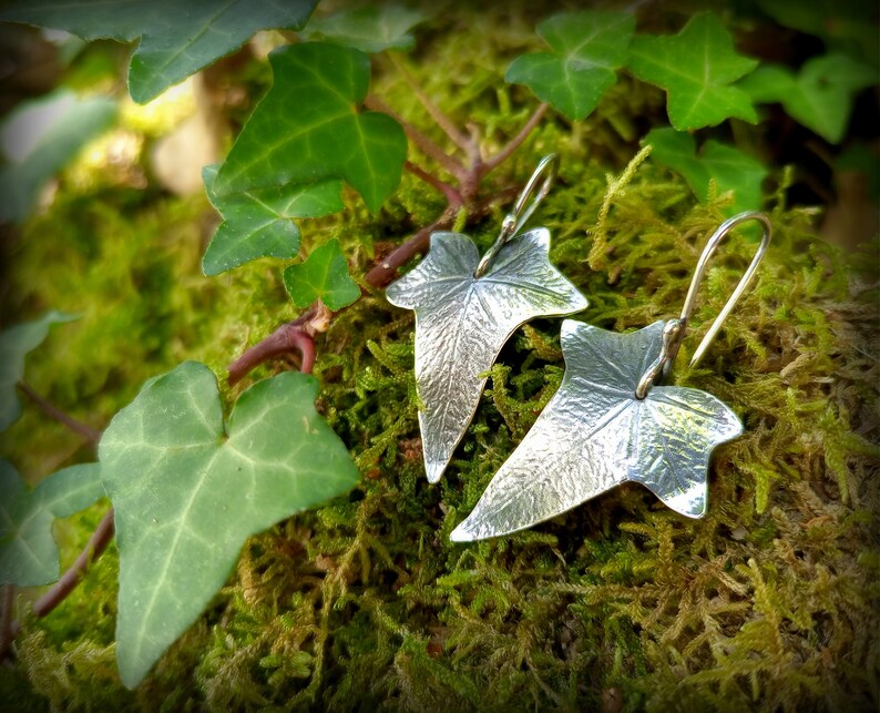 May include: A pair of silver earrings shaped like ivy leaves. The earrings are hanging from a mossy surface.