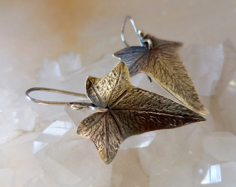 May include: A pair of silver earrings with a textured gold leaf design. The leaves are shaped like ivy leaves and have a detailed, realistic look.