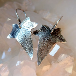 May include: A pair of silver earrings shaped like ivy leaves. The leaves have a textured surface and are detailed with veins.