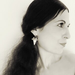 May include: A woman with long dark hair wearing silver leaf earrings. The earrings are shaped like a leaf with a detailed design.