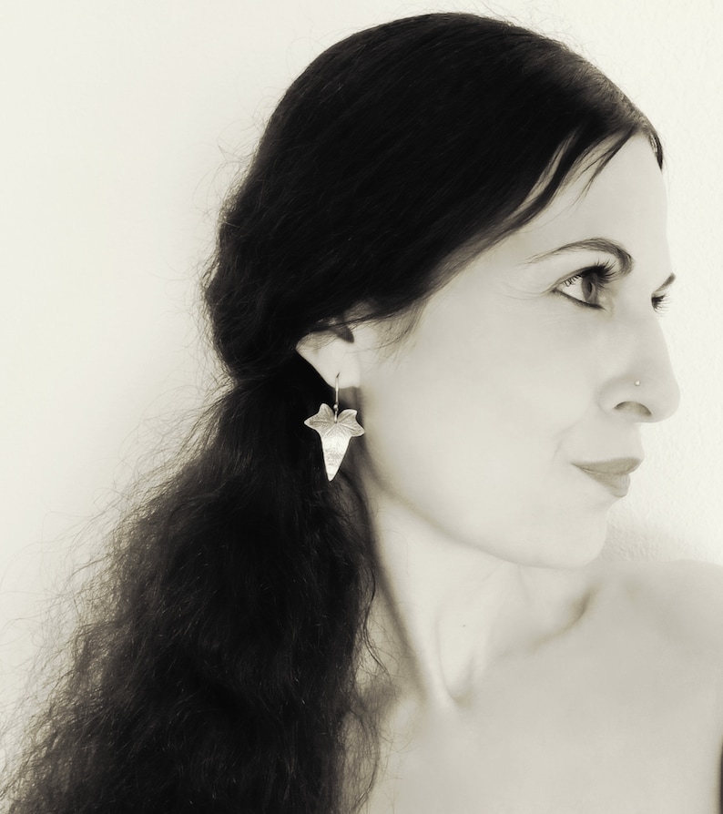 May include: A woman with long dark hair wearing silver leaf earrings. The earrings are shaped like a leaf with a detailed design.