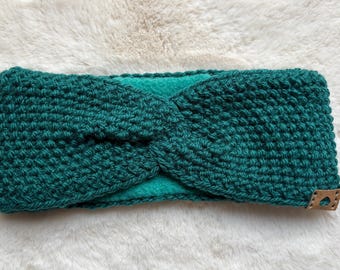Op de afbeelding: Een teal groene gebreide hoofdband met een gedraaid ontwerp. De hoofdband is gemaakt van een getextureerd, dik gebreid materiaal en heeft een zachte teal voering. Een klein bruin leren label is aan de zijkant bevestigd. De hoofdband ligt op een witte, zachte ondergrond.