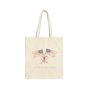 May include: A natural-colored canvas tote bag featuring a watercolor illustration of two American flags tied together with a red ribbon. Below the flags, the text reads LAND OF THE FREE in a simple, dark, sans-serif font centered on the bag.
