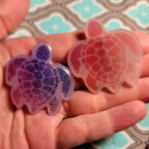 May include: Two resin turtle ornaments, one is pink and the other is purple. Both have glitter embedded in the resin.