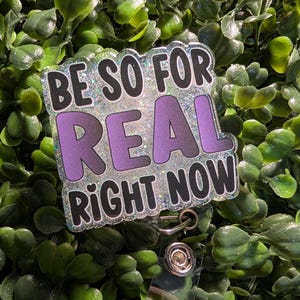 May include: A clear plastic badge holder with a silver glitter background and black text that reads "Be So For Real Right Now". The text "Real" is in purple. The badge holder has a silver metal clip.