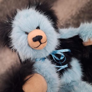 May include: A plush teddy bear featuring long, shaggy fur in alternating patches of black and light blue. The bear has a tan-colored muzzle with a stitched black nose and mouth, dark eyes, and a shiny light blue ribbon tied around its neck.