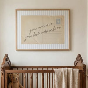 May include: A nursery scene featuring a wooden crib with a teddy bear and a beige blanket. Above the crib hangs a framed print with the text "you are our greatest adventure" on a vintage postcard design.