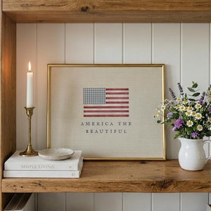 Puede incluir: Una lámina enmarcada con una bandera estadounidense estilizada con un cantón de cuadros azules y blancos y rayas rojas, con el texto AMERICA THE BEAUTIFUL impreso debajo en azul, sobre un estante de madera junto a una vela.