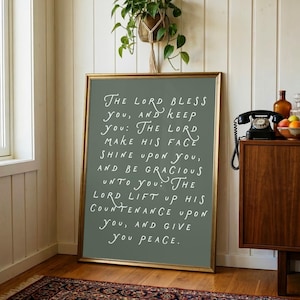 Pode incluir: Uma placa emoldurada com uma borda dourada e um fundo verde sálvia. O texto branco diz: "O Senhor te abençoe e te guarde...e te dê a paz." A placa está encostada em uma parede branca em uma sala com piso de madeira e um tapete estampado.