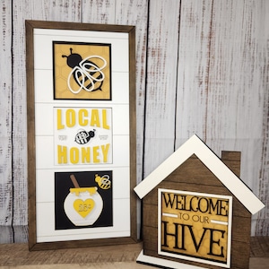 May include: Two wooden signs with a bee theme. The first sign is a white frame with a black bee, honeycomb, and the words "Local Honey". The second sign is a brown house shape with the words "Welcome to our Hive".