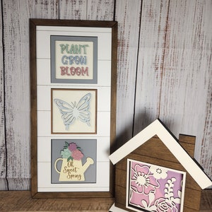 May include: A wooden framed sign with three panels. The top panel says "Plant Grow Bloom" in colorful letters. The middle panel has a blue butterfly cutout. The bottom panel has a yellow watering can with flowers and the text "Oh Sweet Spring". A smaller wooden house shaped sign with a floral cutout is in the foreground.
