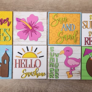 May include: Eight colorful wooden signs with summer-themed text and images. The signs feature phrases like "Summer Vibes", "Sun and Sand", and "Sunrise Sunburn Sunset Repeat".  Other images include a pink hibiscus flower, a brown castle, a yellow sun, a pink flamingo, and a brown coconut with a lime and a purple umbrella.