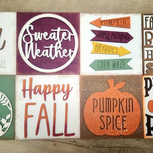 May include: A collection of square wooden signs with fall-themed phrases and images. Signs include "I Love Fall," "Sweater Weather," "Give Thanks," "Happy Fall," and "Pumpkin Spice." Other signs feature pumpkins and fall activities.