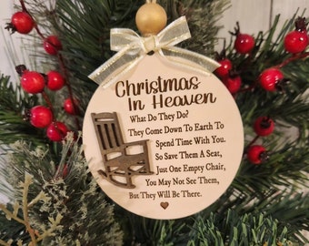 May include: A round, wooden Christmas ornament with the text "Christmas In Heaven" and a poem about loved ones. The ornament features a small rocking chair illustration and is adorned with a gold bow and bead, set against a backdrop of greenery and red berries.