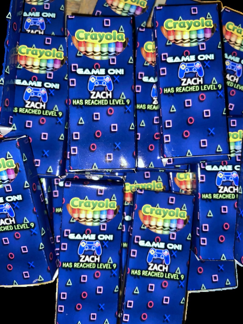 K&ouml;nnte beinhalten: Ein Stapel blauer und schwarzer Crayola-Stifteboxen mit Videospiel-Motiv. Die Boxen haben einen blauen Hintergrund mit einem wei&szlig;en und neongr&uuml;nen Videospiel-Controller und dem Text "GAME ON! ZACH HAS REACHED LEVEL 9".
