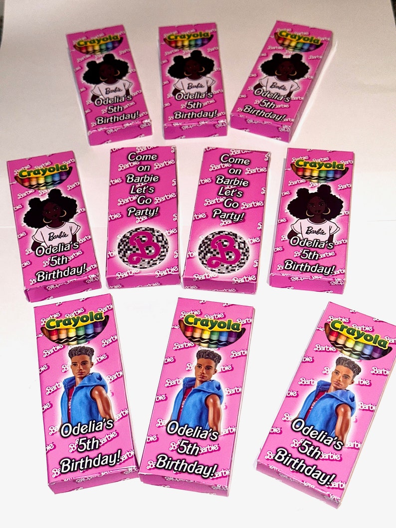 K&ouml;nnte beinhalten: Ein Set aus neun pinkfarbenen und wei&szlig;en Pappkartons mit Crayola-Stiften darin. Jede Schachtel zeigt ein anderes Bild einer Barbie-Puppe auf der Vorderseite. Die Schachteln sind mit dem Text "Come on Barbie Let's Go Party!" und "Odelia's 5th Birthday!" verziert.