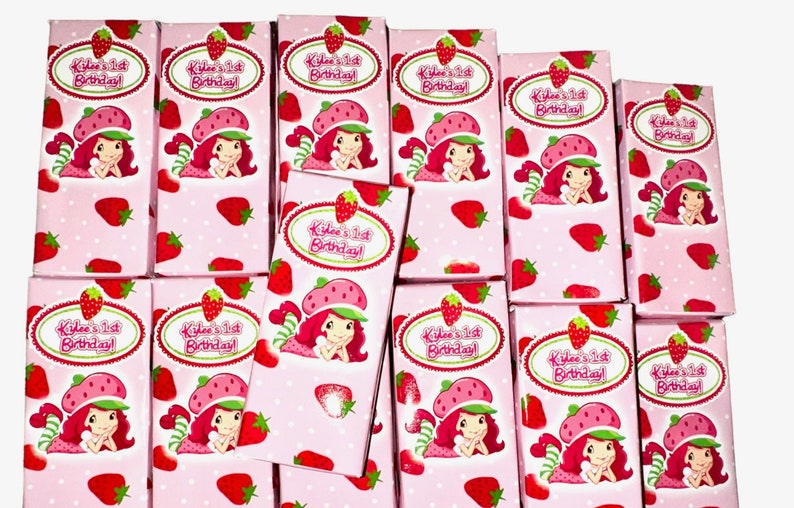 K&ouml;nnte beinhalten: Ein Stapel rosa und wei&szlig;er Geschenkboxen mit einem Erdbeer-Shortcake-Charakterdesign. Die Boxen haben ein wei&szlig;es Oval mit dem Text "Kilee's 1st Birthday!" in gr&uuml;ner und roter Schrift.