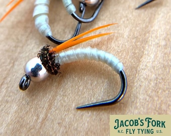 May include: Close-up of three artificial fishing flies. Each fly has a silver bead head, a white body, and orange feather accents. The hooks are black. The flies are arranged on a light brown surface. The text "Jacob's Fork N.C. Fly Tying U.S." is visible.