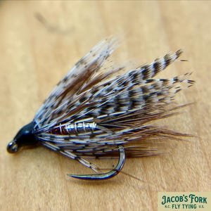 May include: Close-up of a handcrafted fly fishing lure. The lure features a silver hook, black head, and a body wrapped with silver wire. The wings are made of brown and white speckled feathers. The text "JACOB'S FORK N.C. FLY TYING U.S." is visible.