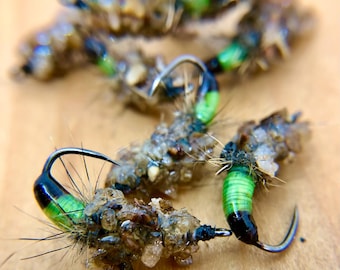 May include: Close-up of several green and black fly fishing lures with brown and white materials. The lures are tied with a hook and have a green and black body. The lures are on a wooden surface. Jacob's Fork N.C. Fly Tying U.S.